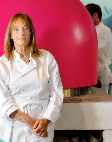 A woman in a chef's uniform stands in front of a bright pink pizza oven with fire burning inside.