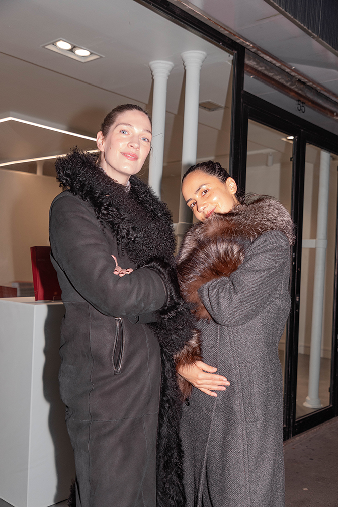 Two women, one in a black fur-trimmed coat and the other smiling in a grey coat with a brown fur collar, stand outside at night.