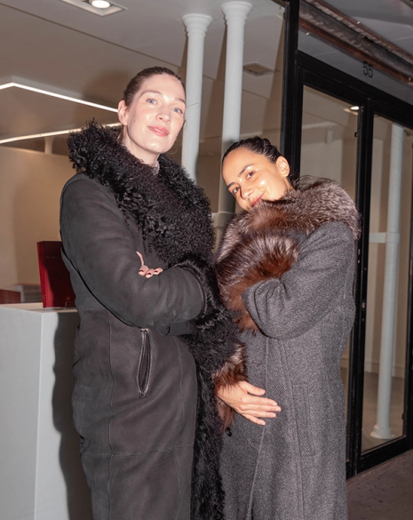 Two women, one in a black fur-trimmed coat and the other smiling in a grey coat with a brown fur collar, stand outside at night.