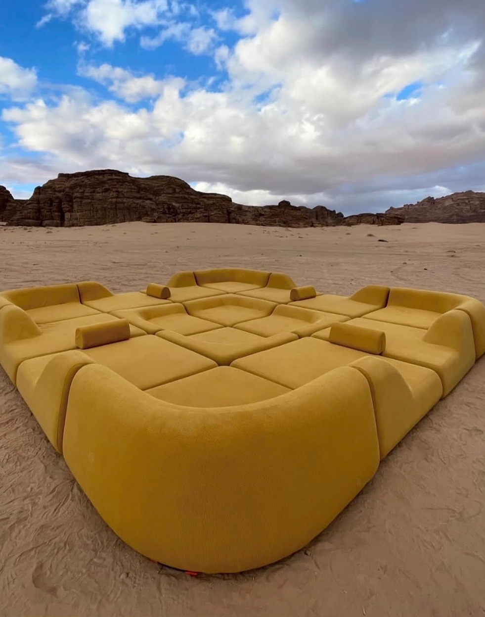 Large modular yellow sofa on sand in a desert with rocky hills and cloudy sky.