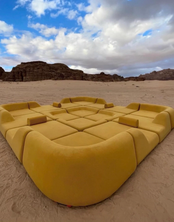 Large modular yellow sofa on sand in a desert with rocky hills and cloudy sky.