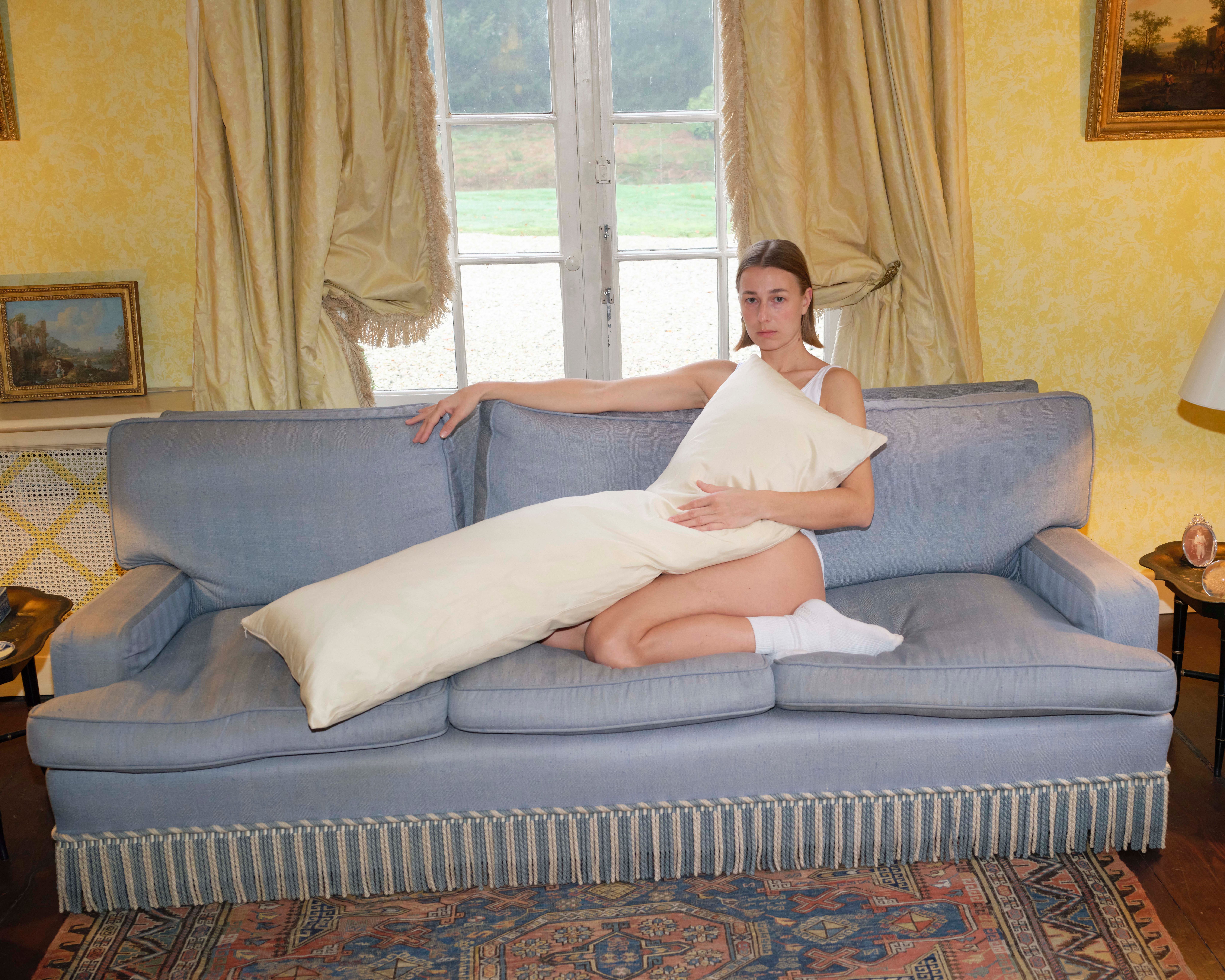 A woman in a white tank top and underwear sits on a blue sofa, holding a long cream-colored pillow across her body.