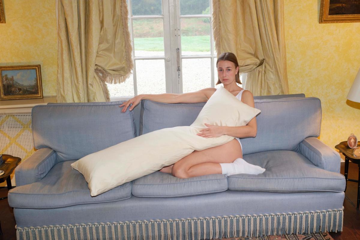 A woman in a white tank top and underwear sits on a blue sofa, holding a long cream-colored pillow across her body.
