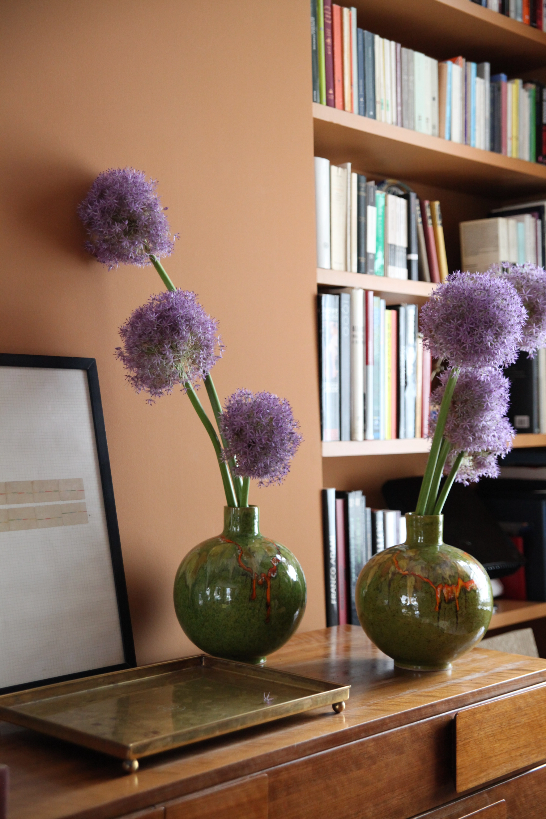Two round green vases filled with purple allium flowers sit on a wooden dresser next to a brass tray, framed artwork, and a bookshelf full of books.