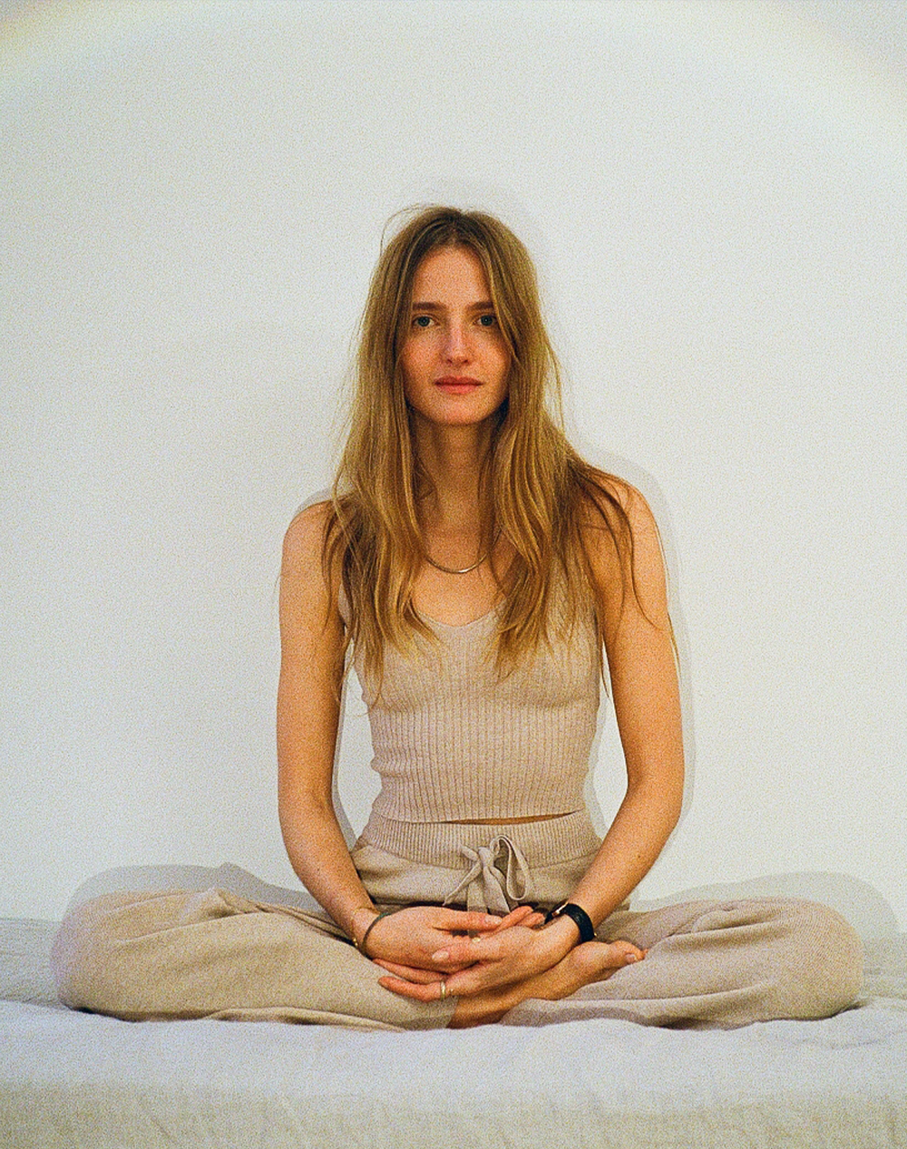 A woman with long blonde hair sits calmly in a meditative cross-legged pose, wearing a beige knit top and matching pants.