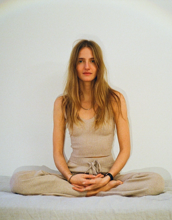 A woman with long blonde hair sits calmly in a meditative cross-legged pose, wearing a beige knit top and matching pants.