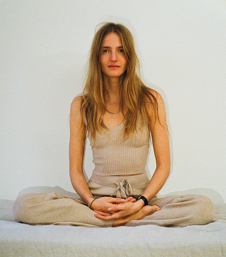 A woman sits cross-legged on a bed wearing a beige tank top and pants, with a subtle rainbow arc on the white wall behind her.