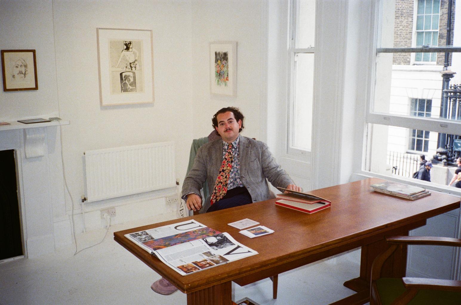 A man with a mustache and a colorful tie sits at a wooden desk in a bright room with artwork on the walls and large windows.