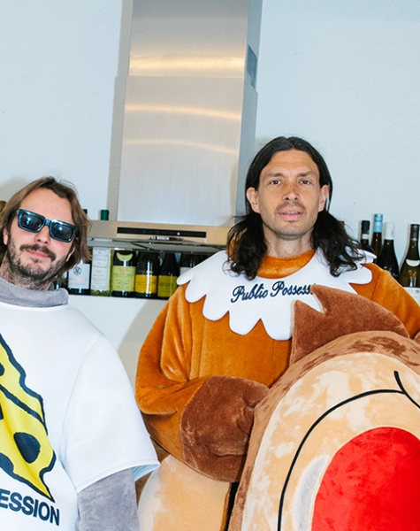 Two men wearing a gray mouse and a brown bear-like plush costume stand in a room with shelves of bottles.