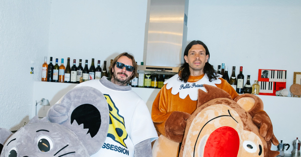 Two men in a gray mouse costume and a brown bear costume stand in a room with bottles on shelves.