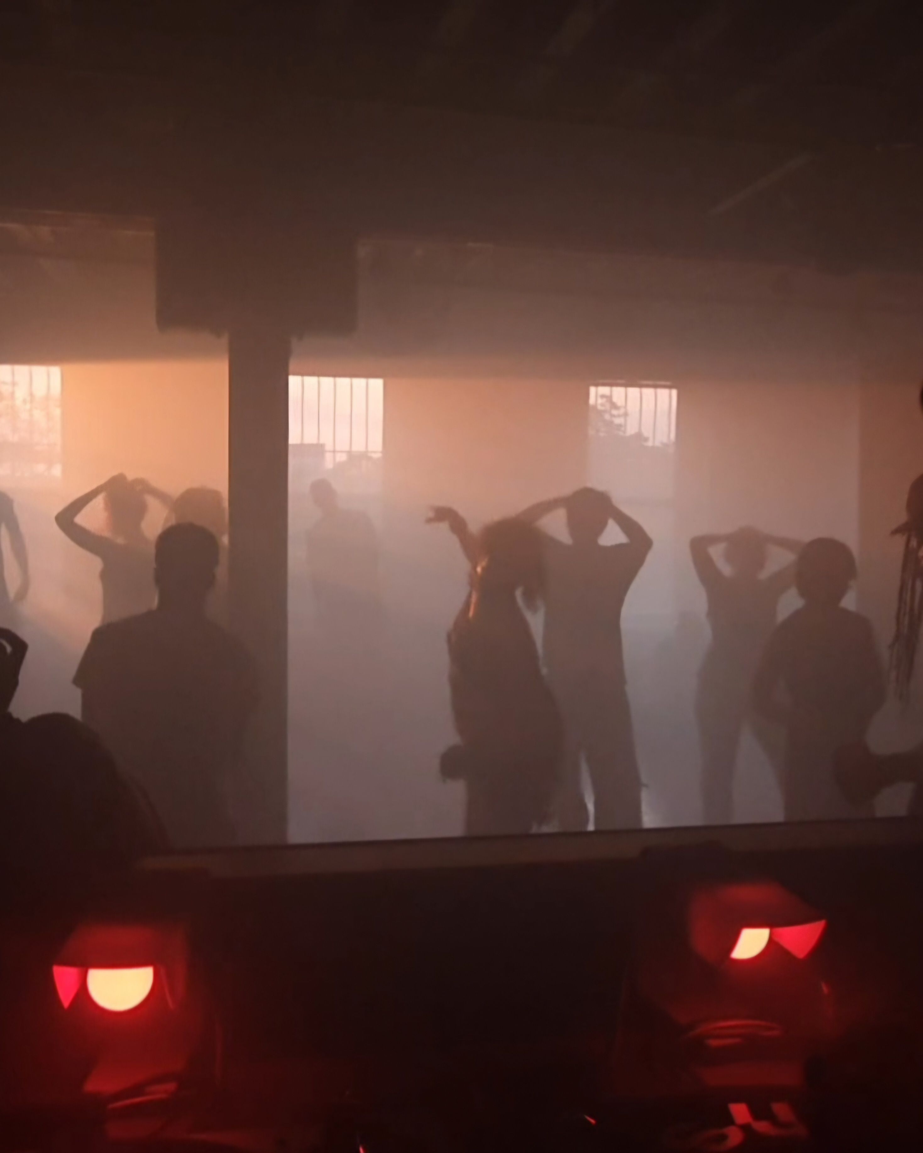 Silhouettes of people dancing in a foggy room lit by an orange glow, with two red lights in the dark foreground.