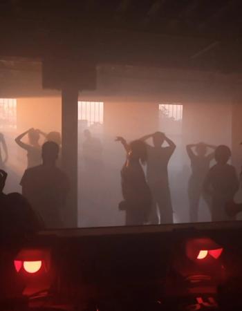Silhouettes of people dancing in a foggy room lit by an orange glow, with two red lights in the dark foreground.