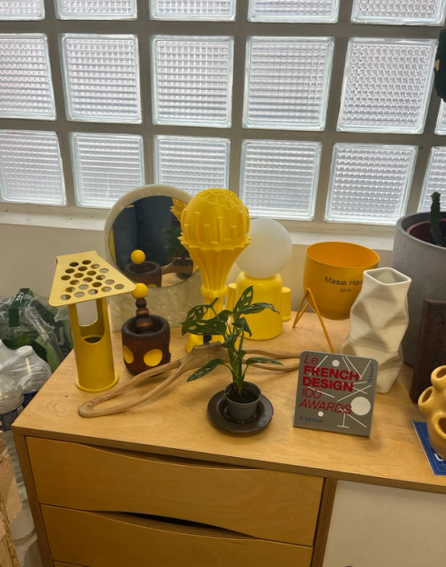 A wooden dresser displays various yellow and white design objects, including lamps and a potted plant, against a glass block window.