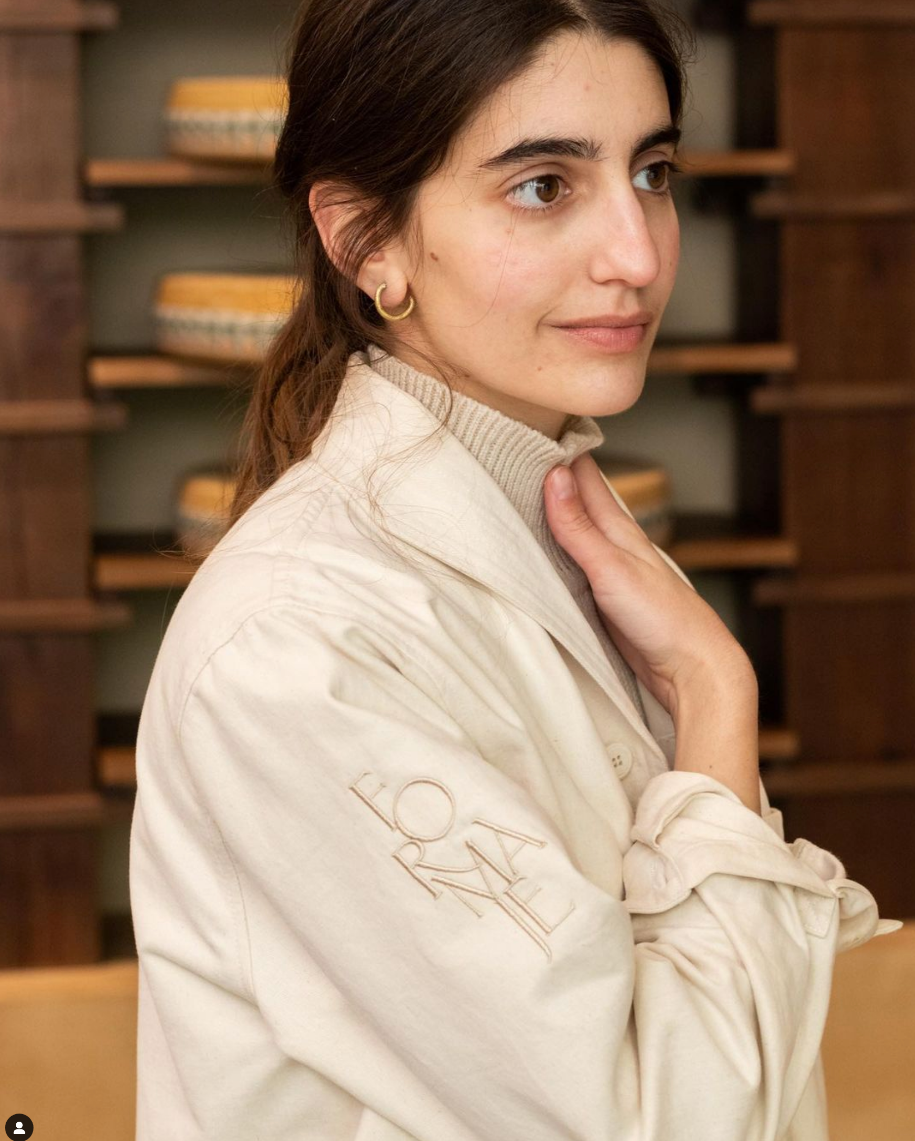 A woman in profile wearing a beige jacket with a "FORMA" logo on the sleeve, her hand near her neck. Shelves with round items are visible behind her.