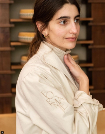 A woman in profile wearing a beige jacket with a "FORMA" logo on the sleeve, her hand near her neck. Shelves with round items are visible behind her.