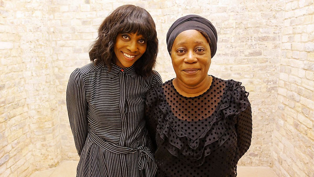 Two smiling Black women, one in a striped dress and the other in a black headwrap, stand against a light brick wall.