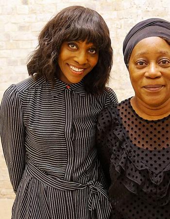 Two smiling Black women, one in a striped dress and the other in a black headwrap, stand against a light brick wall.