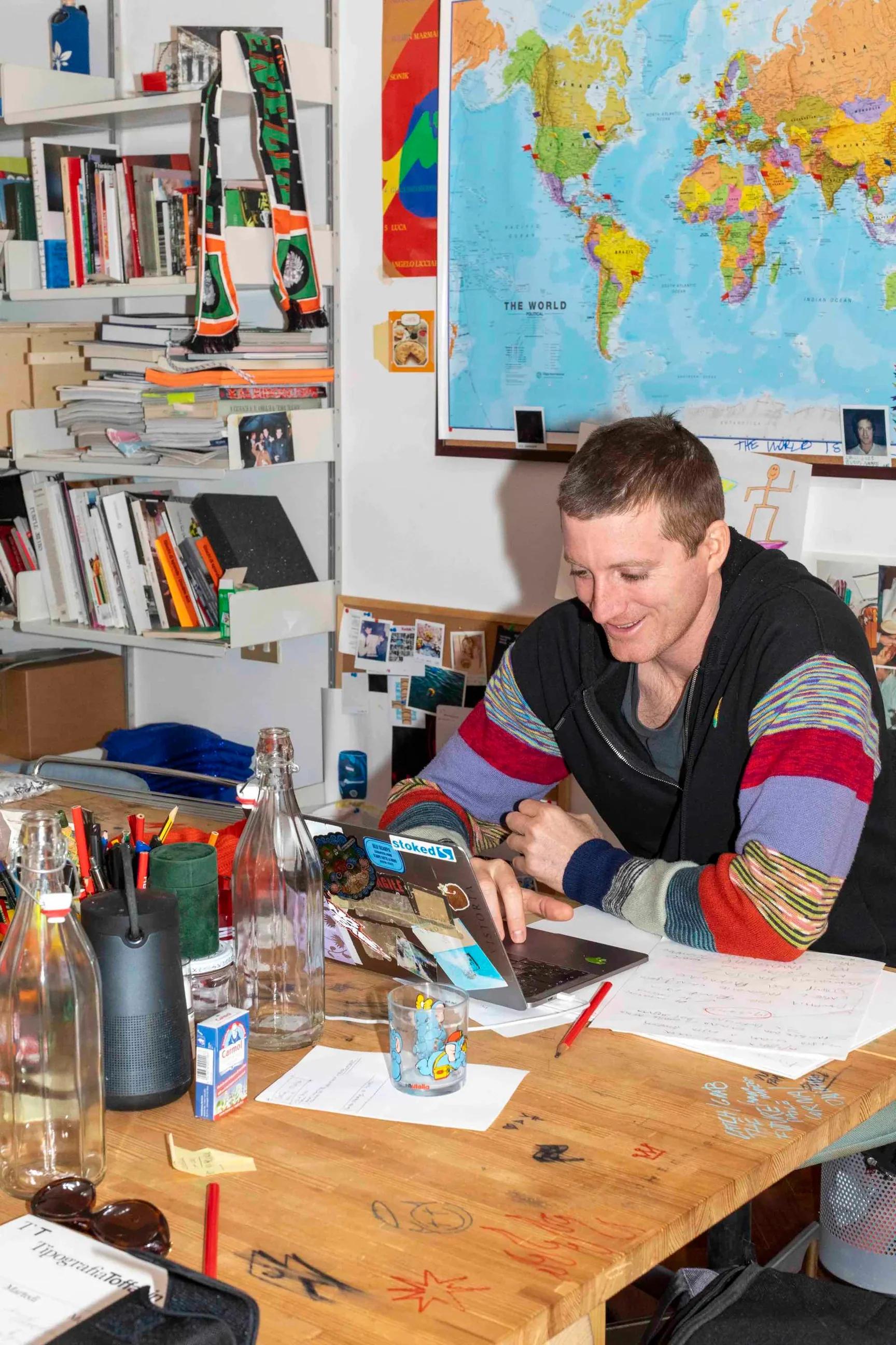 A smiling man in a black jacket with colorful striped sleeves works on a laptop at a cluttered wooden desk with a world map and bookshelves in the background.
