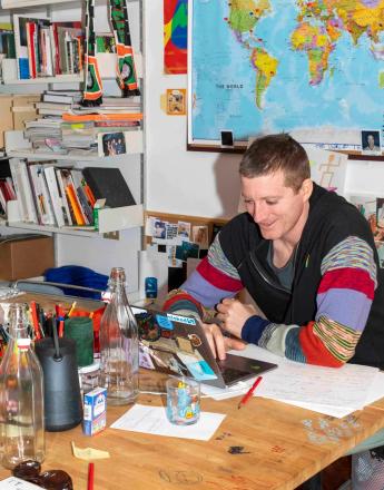 A smiling man in a black jacket with colorful striped sleeves works on a laptop at a cluttered wooden desk with a world map and bookshelves in the background.