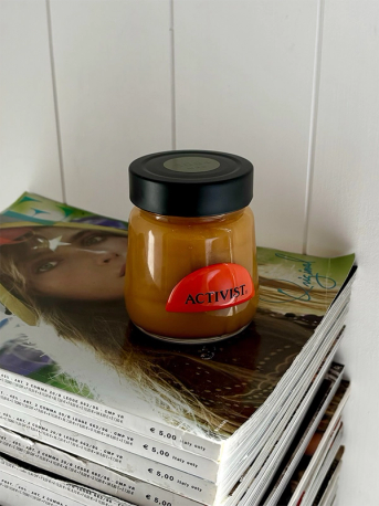 A glass jar of amber-colored spread with a red "ACTIVIST" label rests on a stack of magazines.