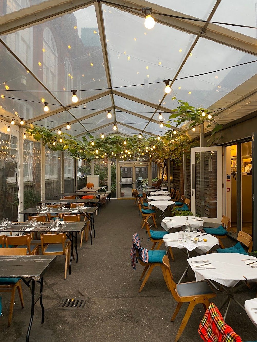 A covered outdoor restaurant patio with string lights, green vines, and many tables set for dining.