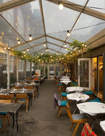 A covered outdoor restaurant patio with string lights, green vines, and many tables set for dining.