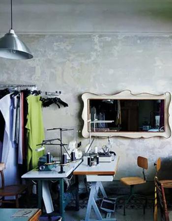 A sewing studio with two machines, a clothing rack, and a decorative mirror on a textured grey wall.