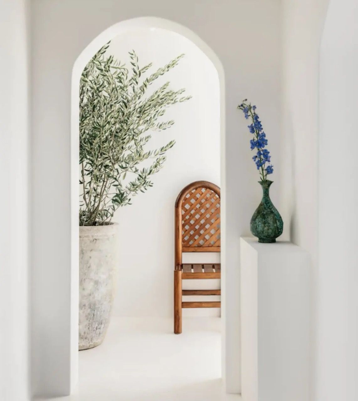 Minimalist white interior with an arched opening, a potted olive tree, wooden chair, and a green vase with blue flowers on a white pedestal.