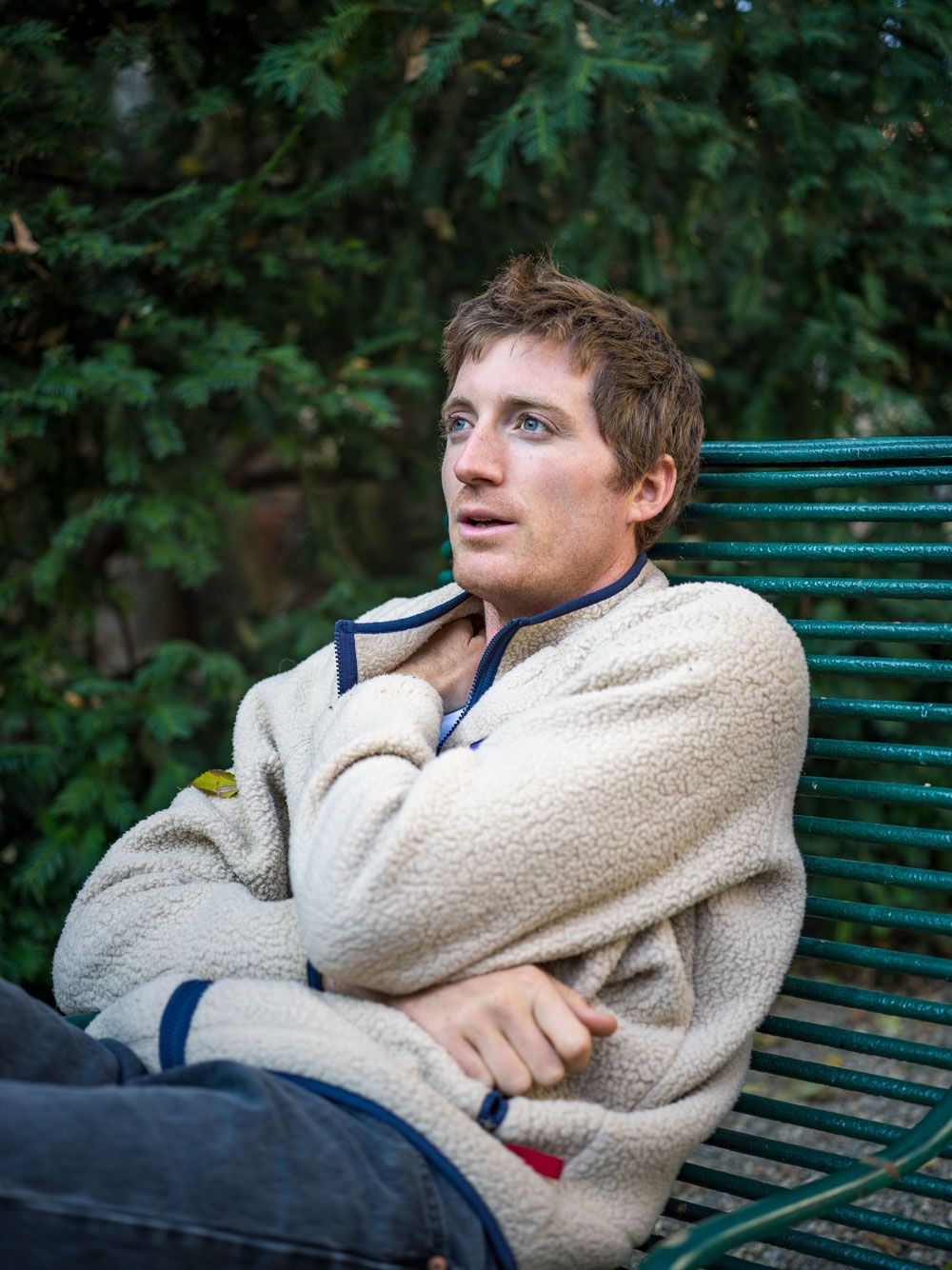 A young man in a cream fleece jacket sits on a green bench, looking thoughtfully upwards.