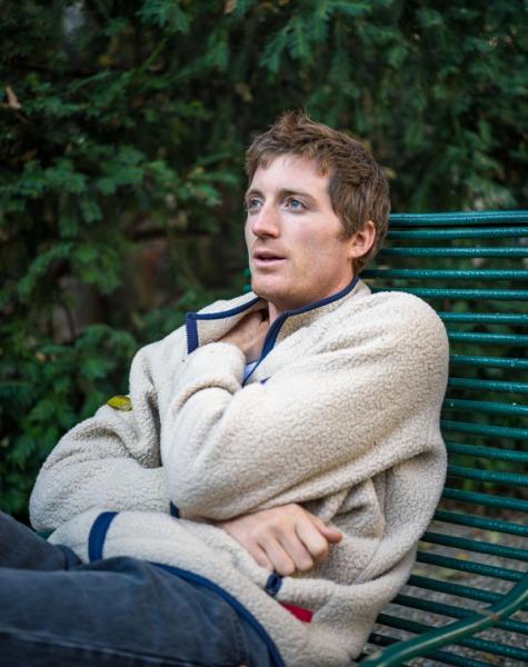 A young man in a cream fleece jacket sits on a green bench, looking thoughtfully upwards.