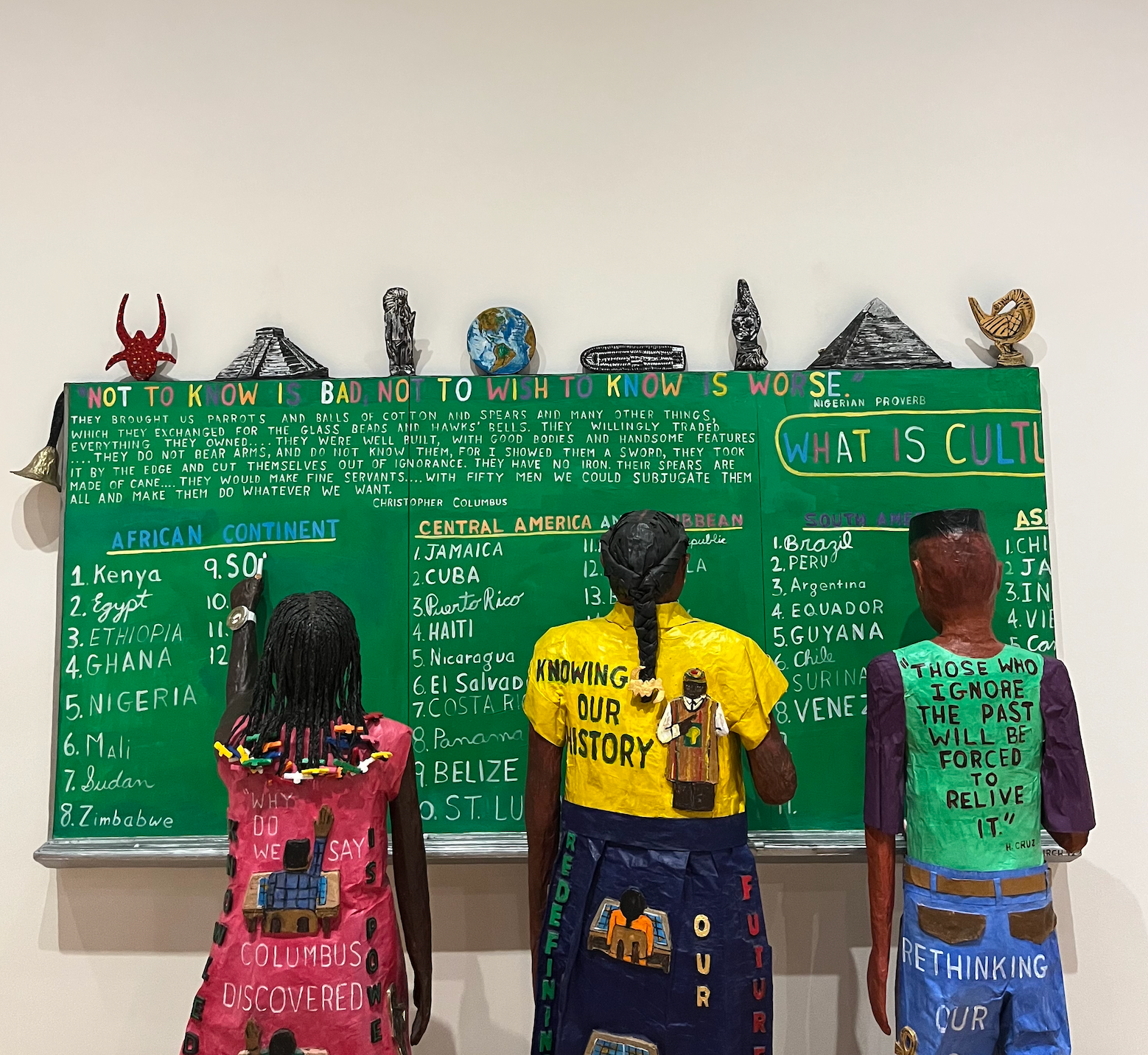 An art installation featuring three sculptural figures facing a green chalkboard covered with historical text, country lists, and questions about culture, with messages about history and knowledge on their clothing.