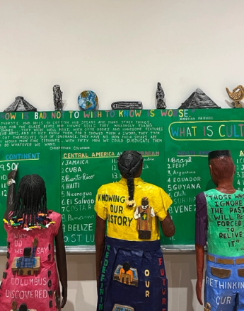 An art installation featuring three sculptural figures facing a green chalkboard covered with historical text, country lists, and questions about culture, with messages about history and knowledge on their clothing.