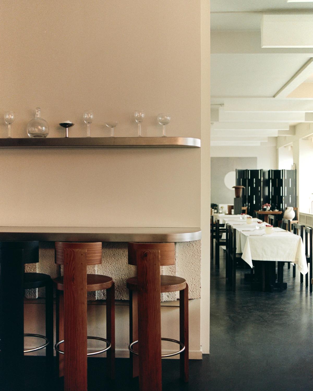 Modern restaurant interior with a bar counter and wooden stools on the left, and a dining room with white-clothed tables on the right.