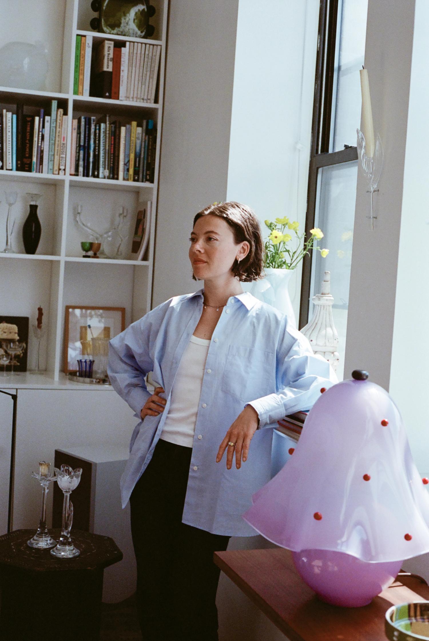 A woman in a light blue shirt stands in a brightly lit room, a white bookshelf behind her and a purple mushroom lamp beside her.
