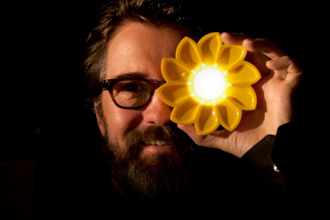 A smiling man with a beard and glasses holds a bright yellow flower-shaped light over his left eye in the dark.