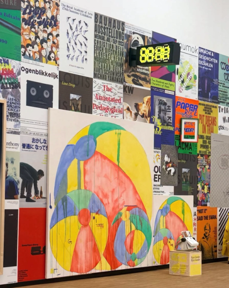A gallery wall is covered in numerous exhibition posters, with a large painting of dripping red, yellow, and blue spheres in the foreground, and a digital clock displaying 88:88.