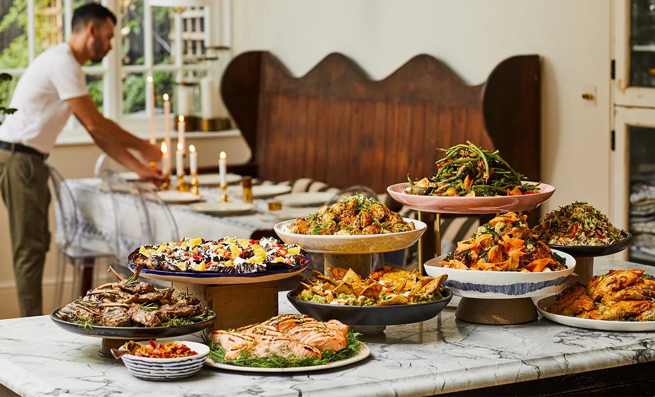 A lavish spread of various dishes, including salmon, lamb chops, and salads, on a marble table, with a man setting a dining table in the background.