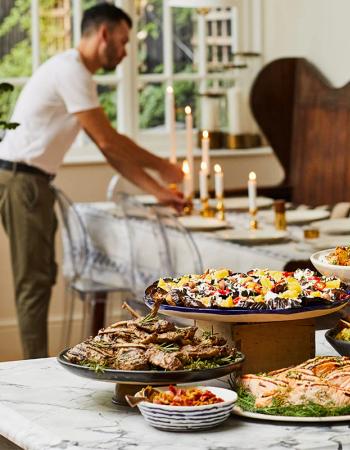 A lavish spread of various dishes, including salmon, lamb chops, and salads, on a marble table, with a man setting a dining table in the background.