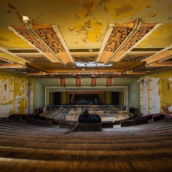 Abandoned theater with peeling yellow paint, intricate decaying ceiling, and rows of worn wooden seats facing a dilapidated stage.
