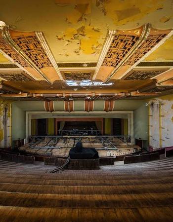 Abandoned theater with peeling yellow paint, intricate decaying ceiling, and rows of worn wooden seats facing a dilapidated stage.