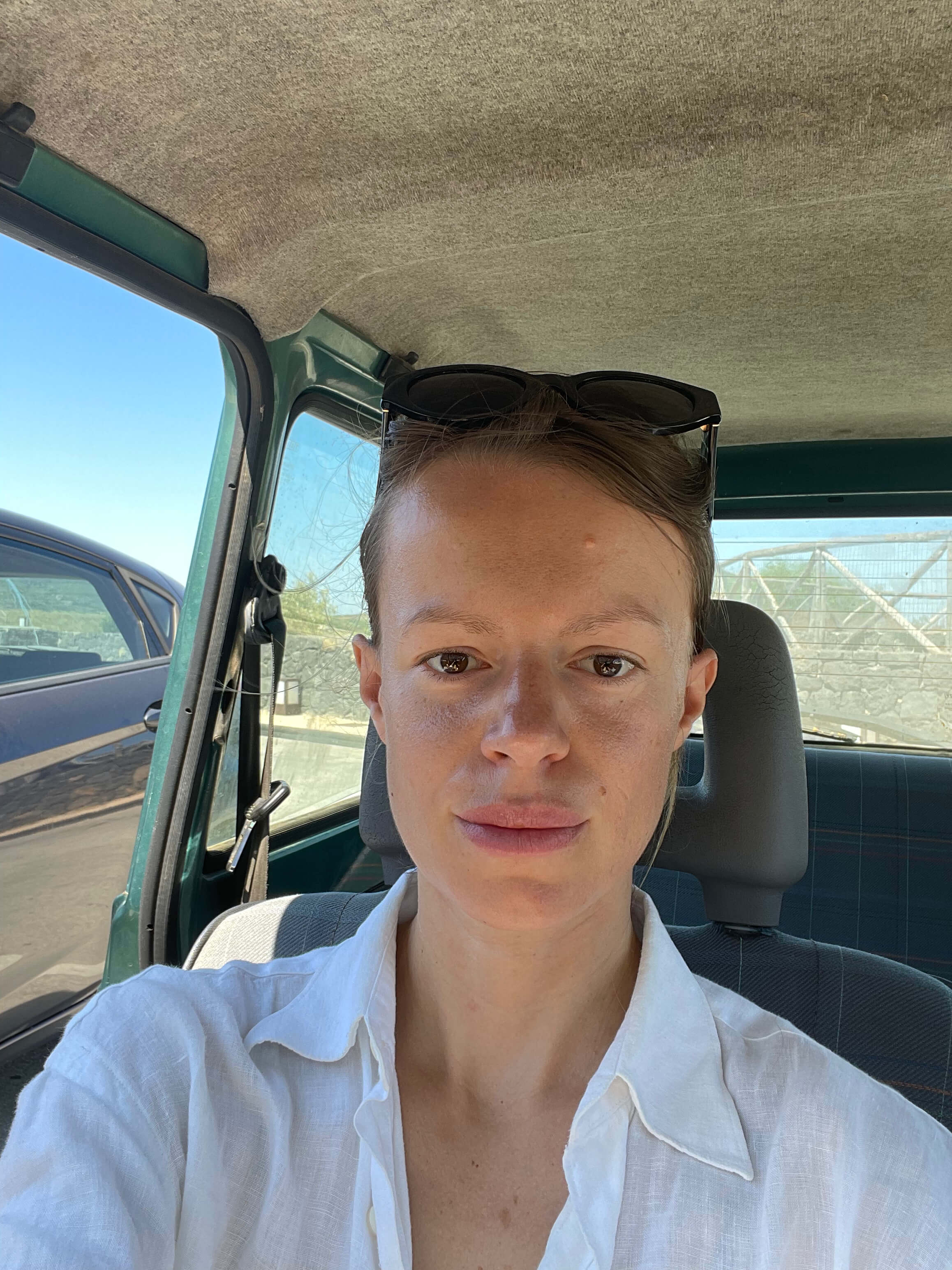 A woman in a white shirt with sunglasses on her head, sitting in a car.