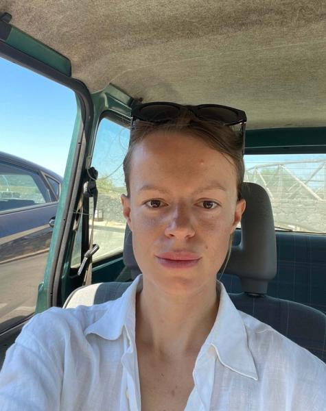 A woman with sunglasses on her head, wearing a white shirt, takes a selfie inside a car.