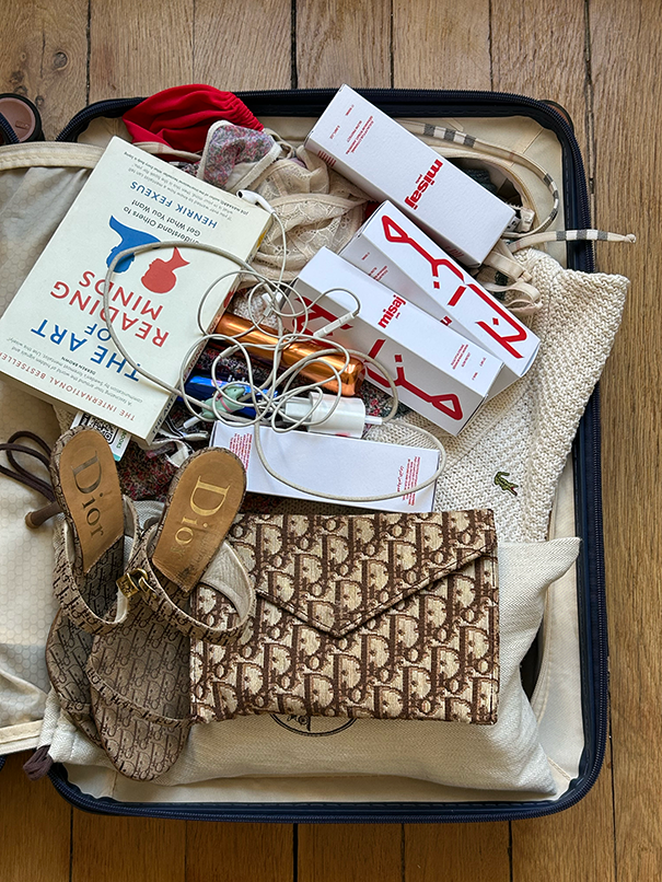An open suitcase packed with a book, Dior sandals and clutch, Misaj products, a Lacoste sweater, and other clothes.