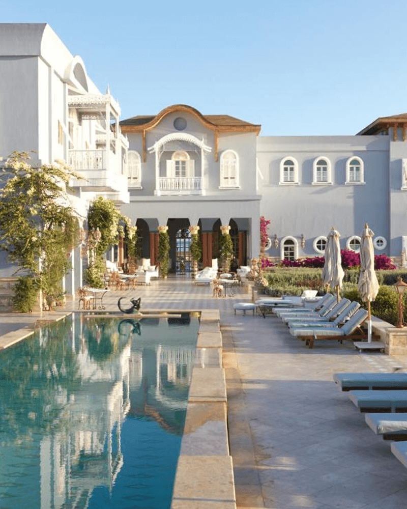 Elegant resort with a long swimming pool, lounge chairs, and a light-blue building under a clear sky.