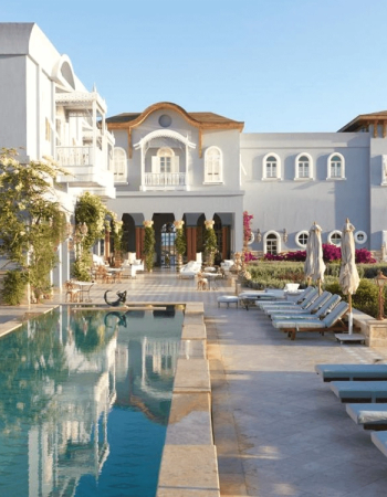 Elegant resort with a long swimming pool, lounge chairs, and a light-blue building under a clear sky.