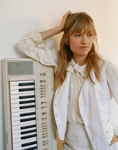 A woman with blonde hair and bangs leans against a vintage Yamaha keyboard with her hand on her head.