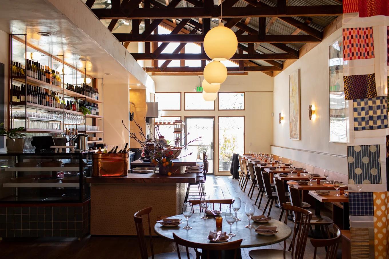 Restaurant interior with a bar, set dining tables, exposed wooden beams, pendant lights, and decorative fabric panels.