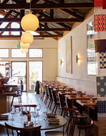 Restaurant interior with a bar, set dining tables, exposed wooden beams, pendant lights, and decorative fabric panels.