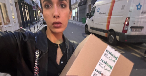 A person in a black jacket holds a brown package on a city street, with a white delivery van in the background.