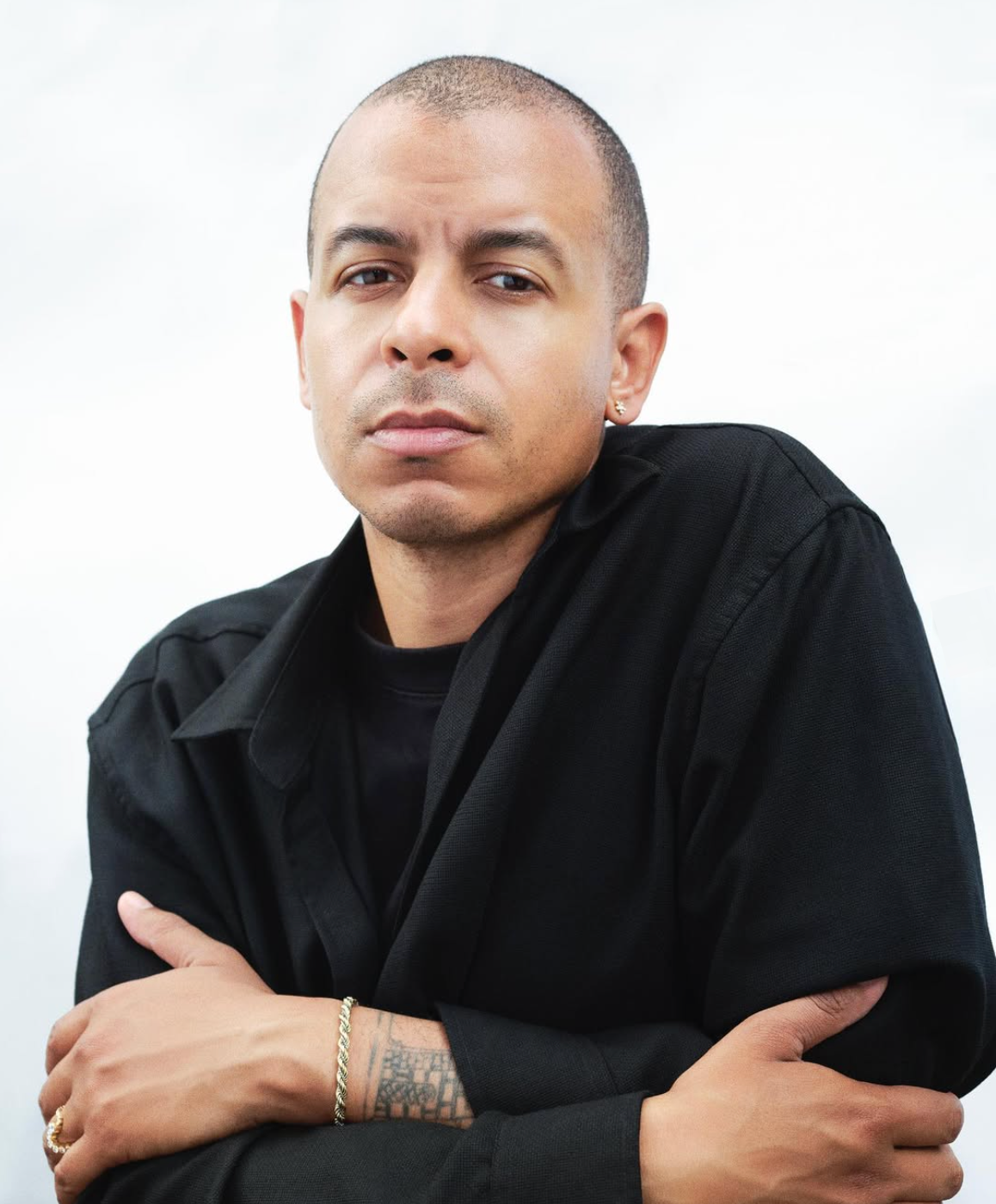 A man with a shaved head and light brown skin, wearing a black shirt, crosses his arms and looks seriously at the camera. He wears a gold earring and a gold bracelet.